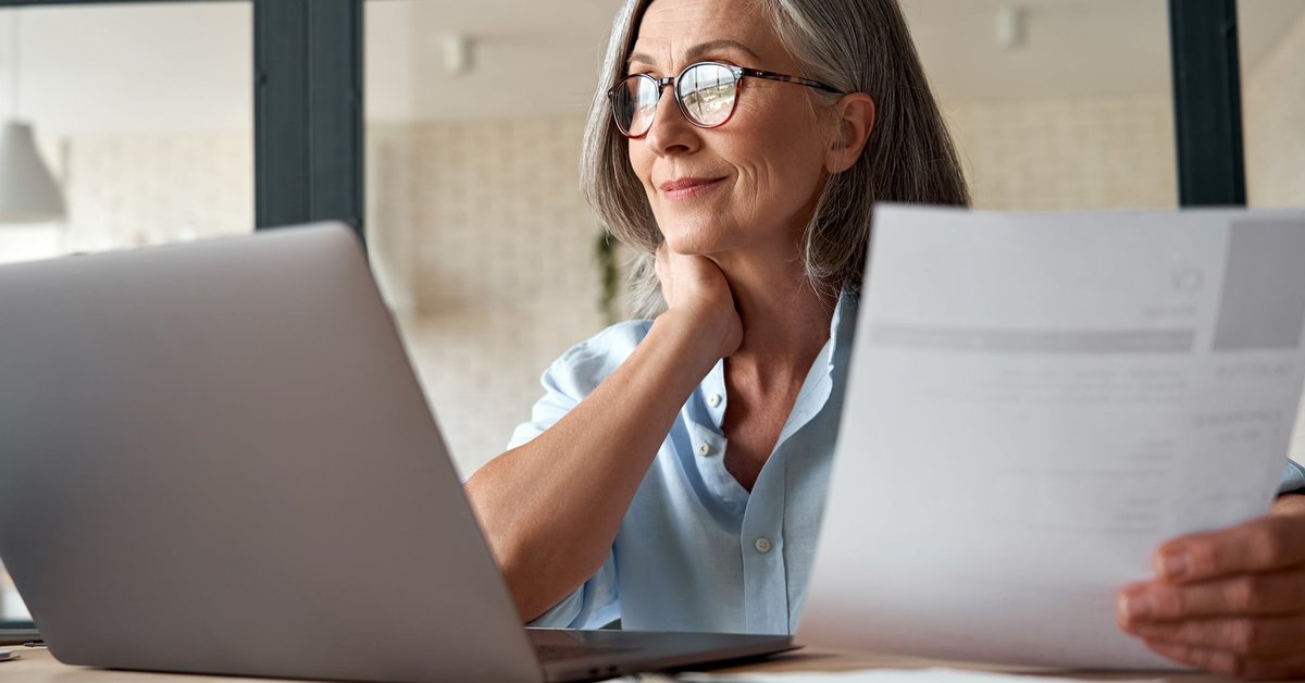 Mature woman working from laptop with documents in hand.