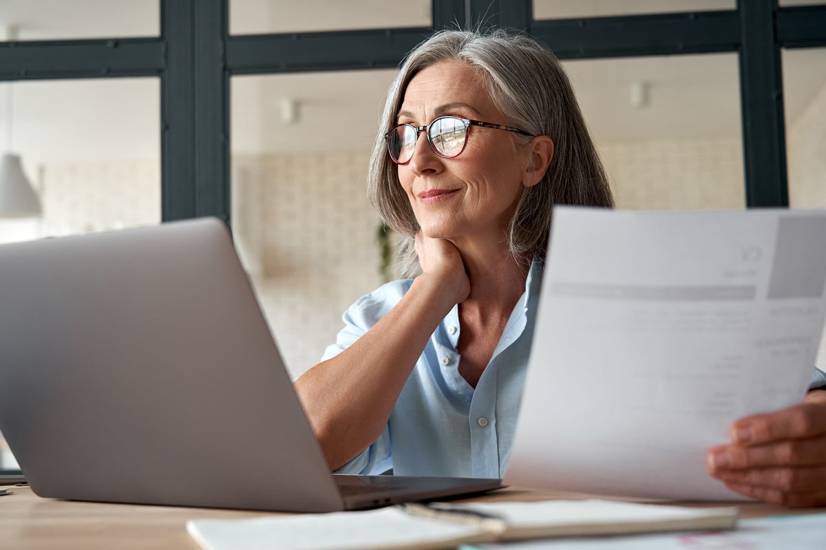 Mature woman working from laptop with documents in hand.