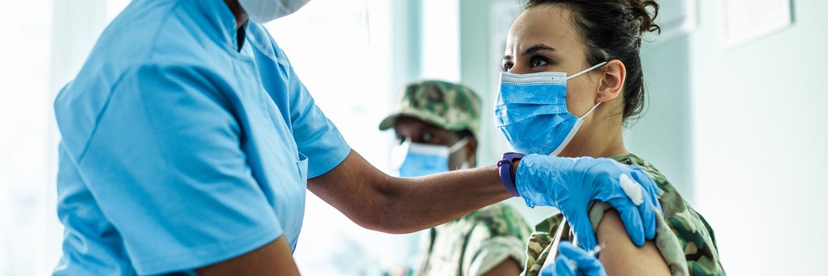 A medical staff member in a face shield provides an injection to a patient wearing a mask.