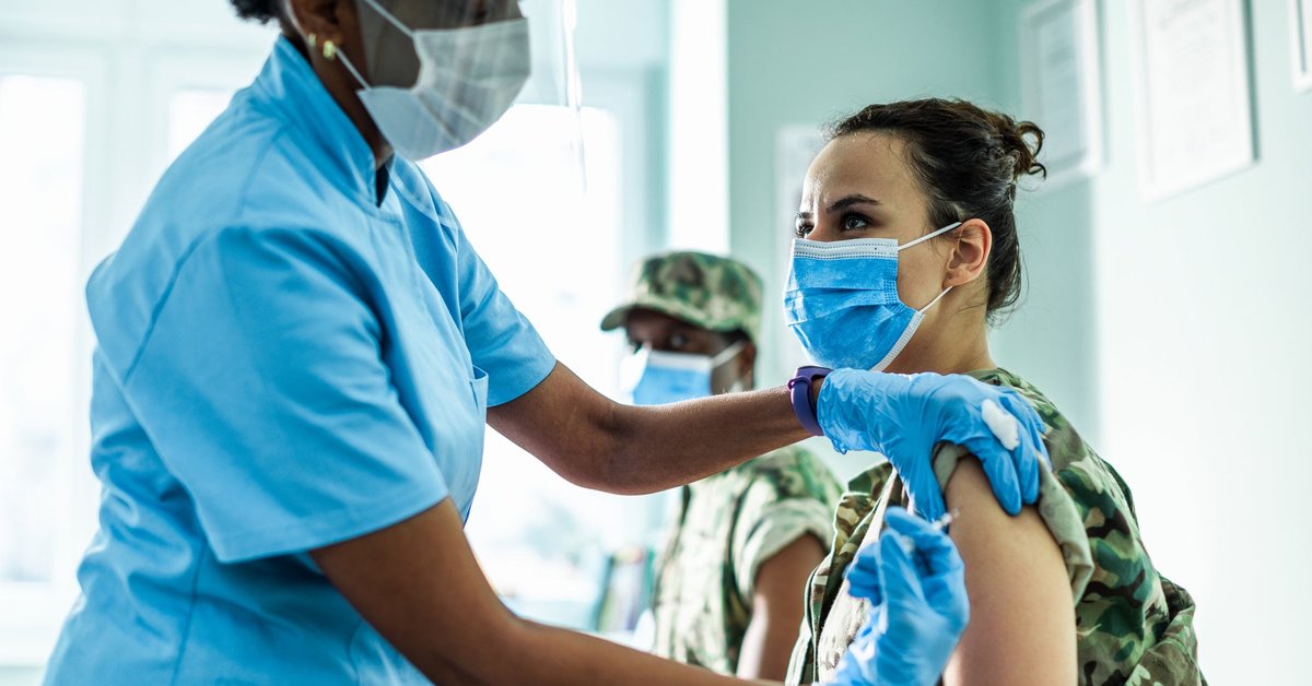 A medical staff member in a face shield provides an injection to a patient wearing a mask.