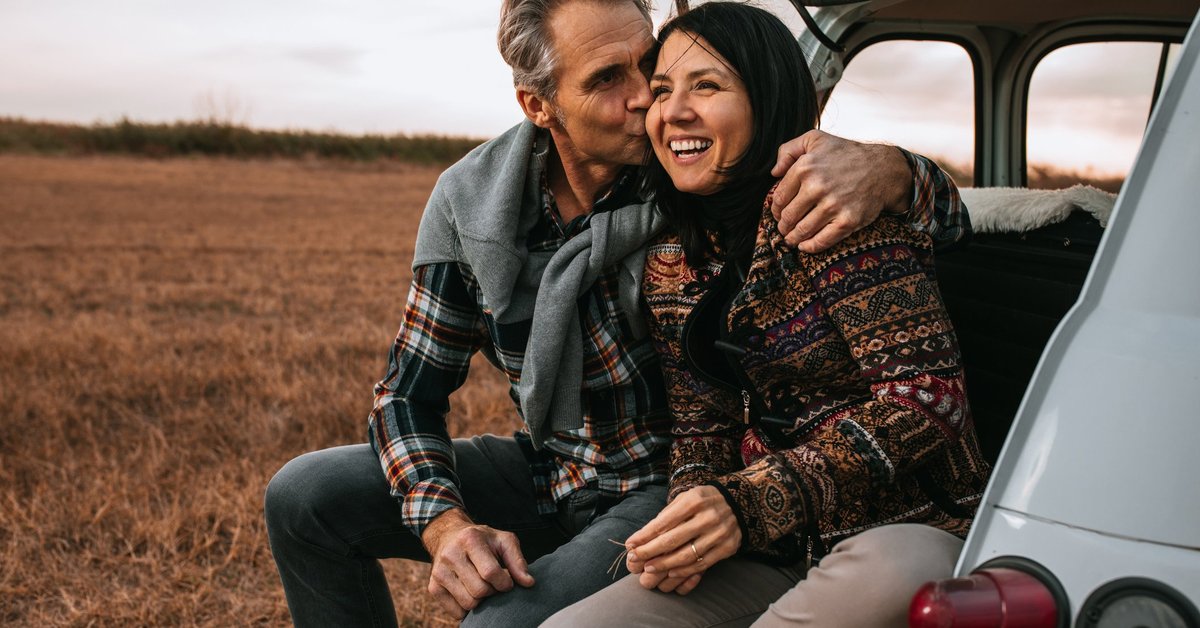 A middle-age couple smiling and sitting on the back of a car in a field.