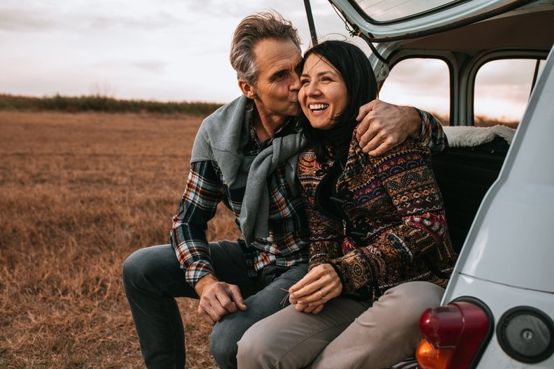 A middle-aged couple sitting in the open trunk of their car in a field with their arms around each other.
