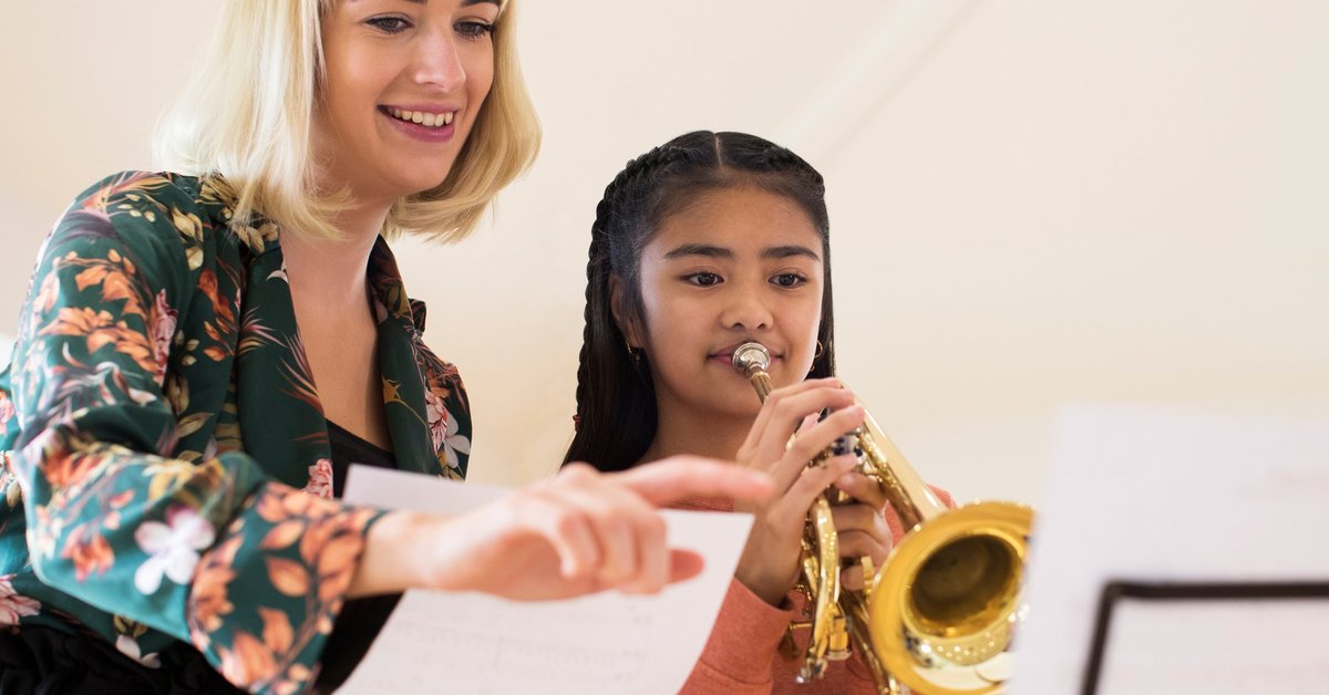 A smiling female teacher instructing a young girl playing a trumpet.