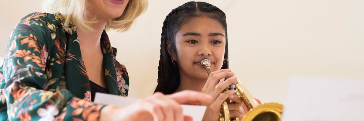 A smiling female teacher instructing a young girl playing a trumpet.