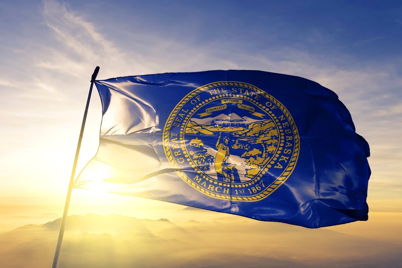 The Nebraska state flag flying in front of a sunny sky.