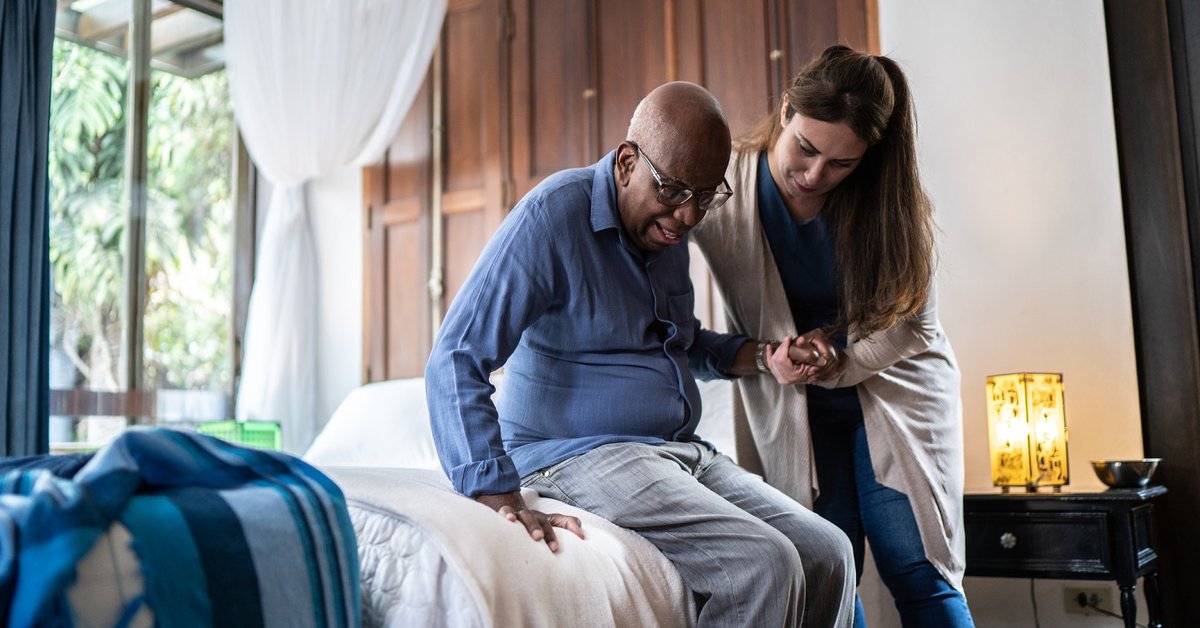 Nurse helping elderly man from bed in nursing home.