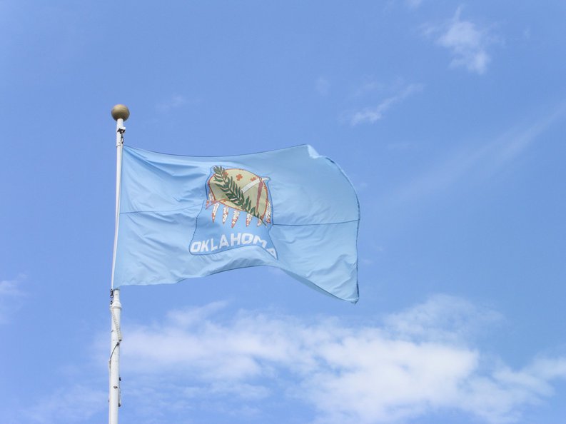 The Oklahoma state flag in front of a blue sky and white clouds.