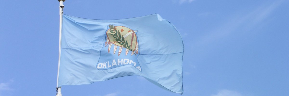 The Oklahoma state flag in front of a blue sky and white clouds.
