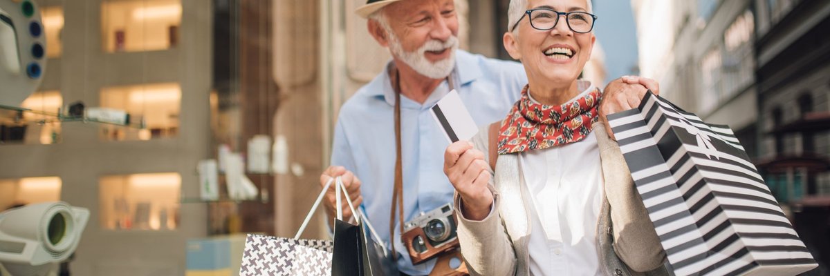 An older couple, out on a credit card spending spree, are shown walking down a street, holding shopping bags and smiling.