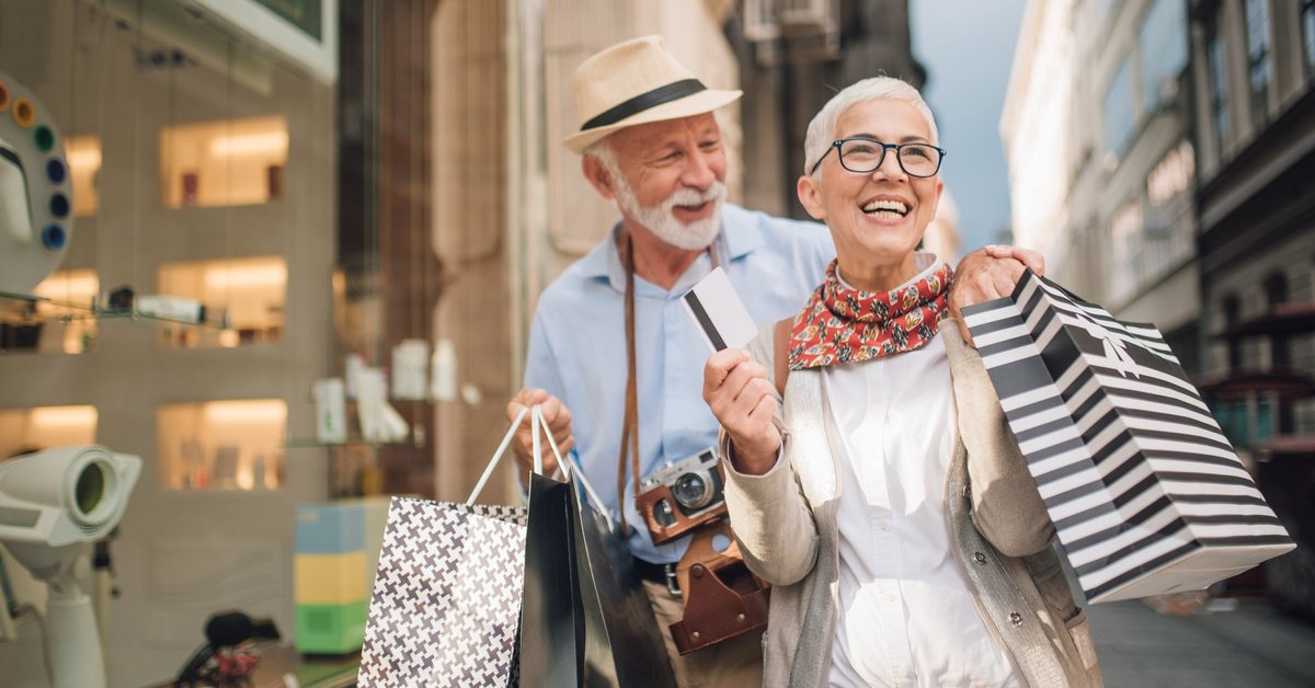 An older couple, out on a credit card spending spree, are shown walking down a street, holding shopping bags and smiling.