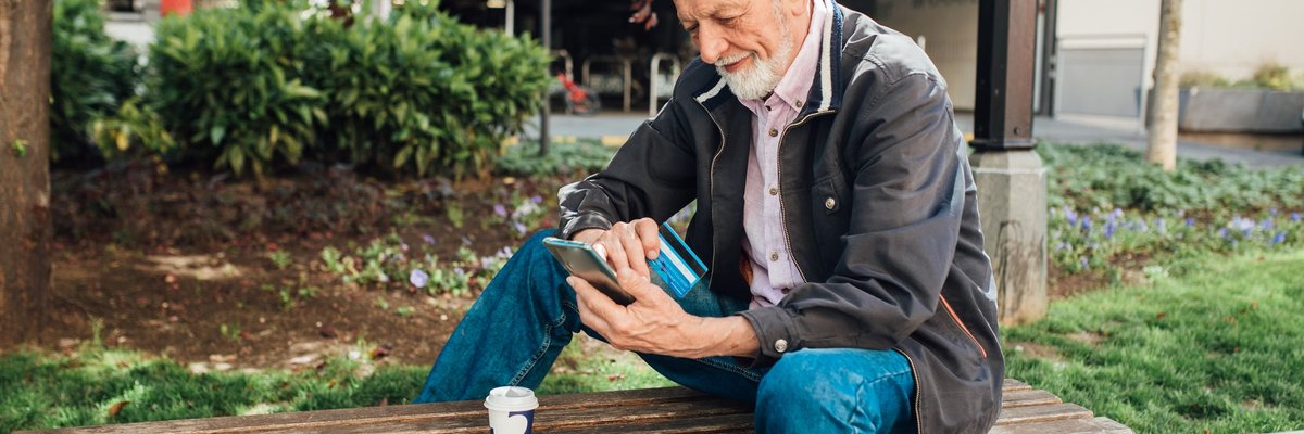 An older gentleman sits on a park bench, looking at his phone while also holding his credit card.