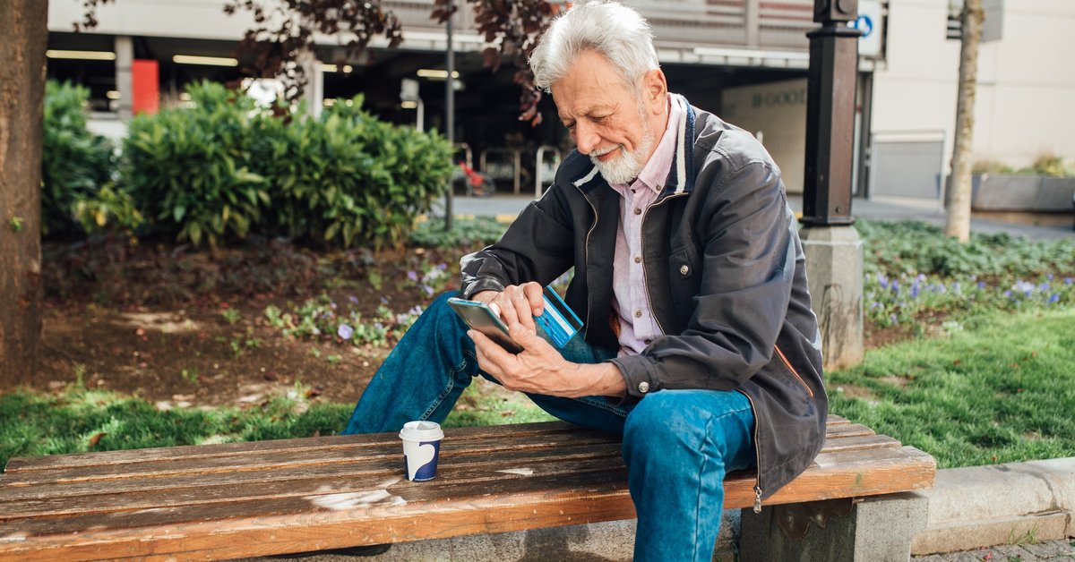 An older gentleman sits on a park bench, looking at his phone while also holding his credit card.