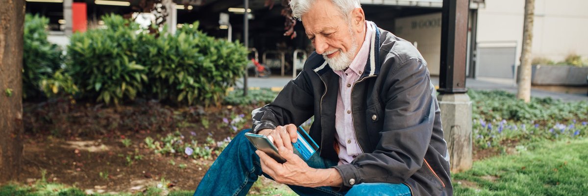 An older gentleman sits on a park bench, looking at his phone while also holding his credit card.