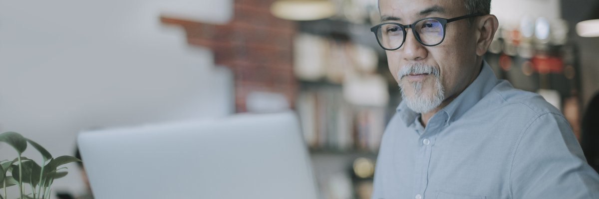 An older man uses his laptop to read the news while sitting in a coffee shop.