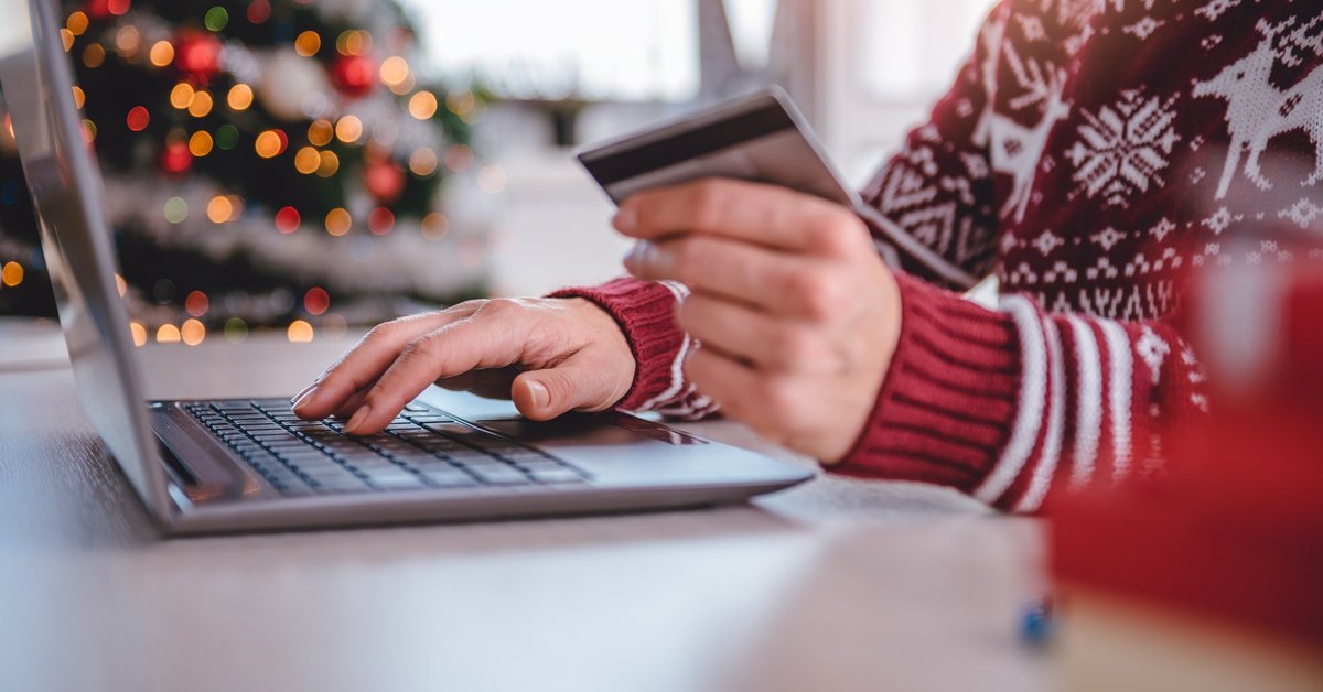 A person in a holiday sweater holding a credit card while typing on a laptop.