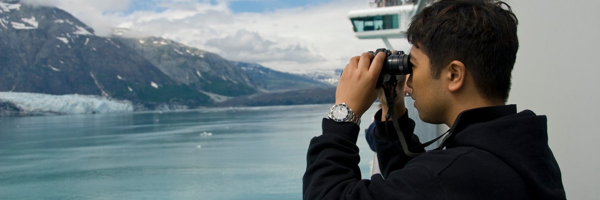 A person looking through binoculars at snowy mountains from the deck of a cruise ship.