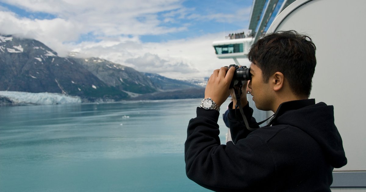 A person looking through binoculars at snowy mountains from the deck of a cruise ship.