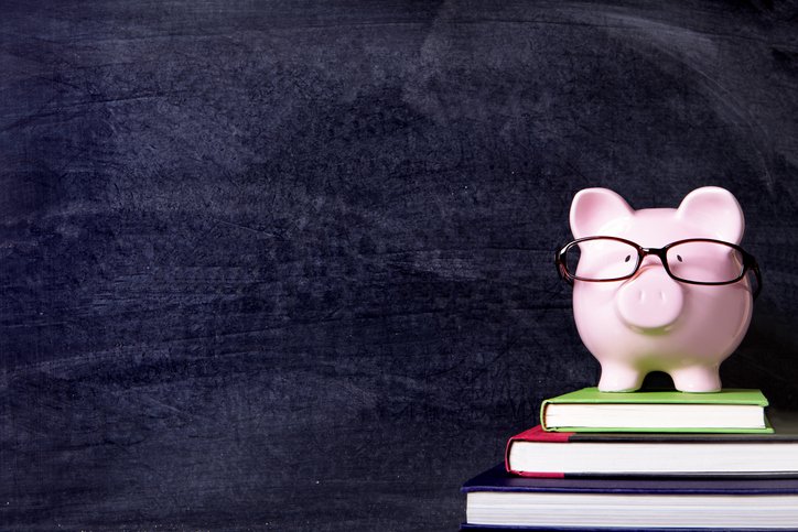 Piggy bank wearing glasses sitting on stack of books in front of classroom chalkboard.
