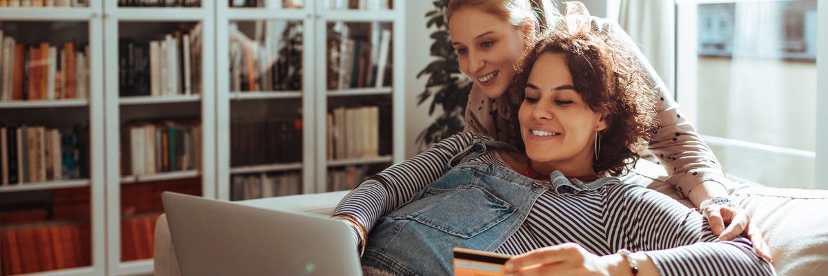 Pregnant woman holding credit card while using laptop with another woman looking over her shoulder.