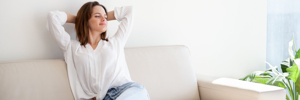 A relaxed woman sitting on her couch with her hands behind her head.