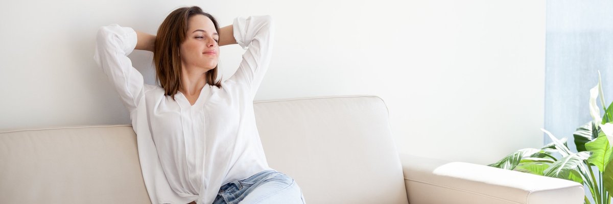 A relaxed woman stretching her arms above her head while sitting on her couch in a sunny apartment.