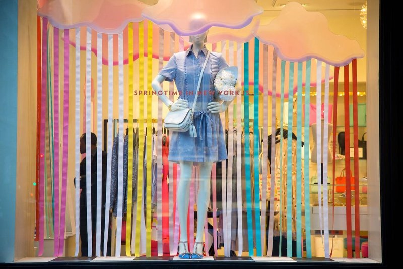 Mannequin standing against multicolored strips of paper and pink clouds in a store window.