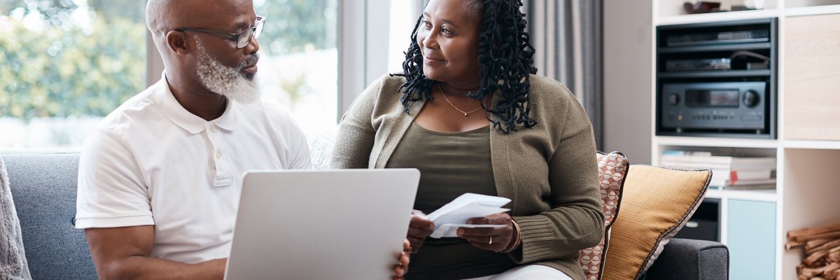Senior couple sitting on couch together going over information on a laptop and other documents.
