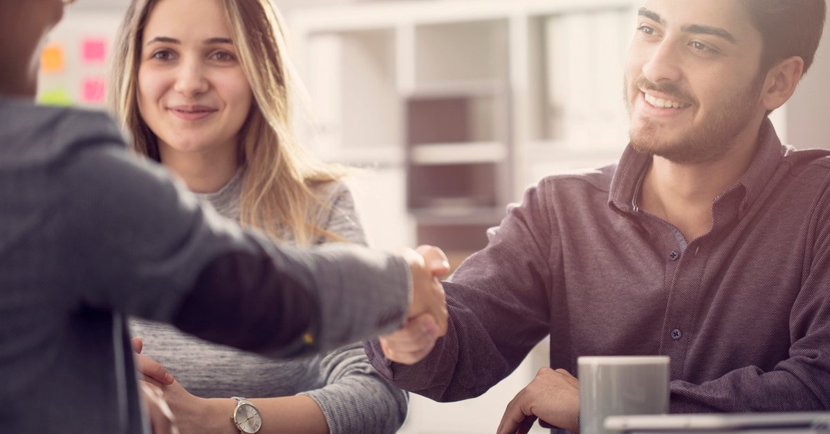 A smiling young couple shaking hands with someone over a desk.