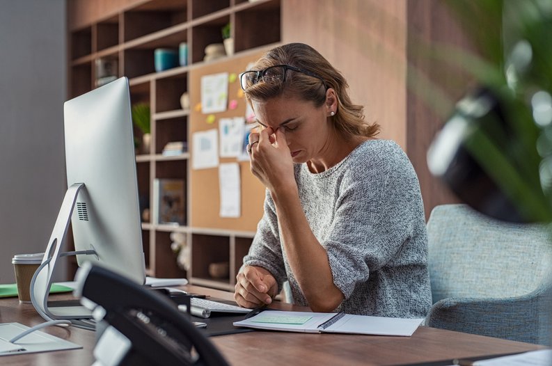 A stressed woman sitting at her office desk with her fingers pinching the bridge of her nose.