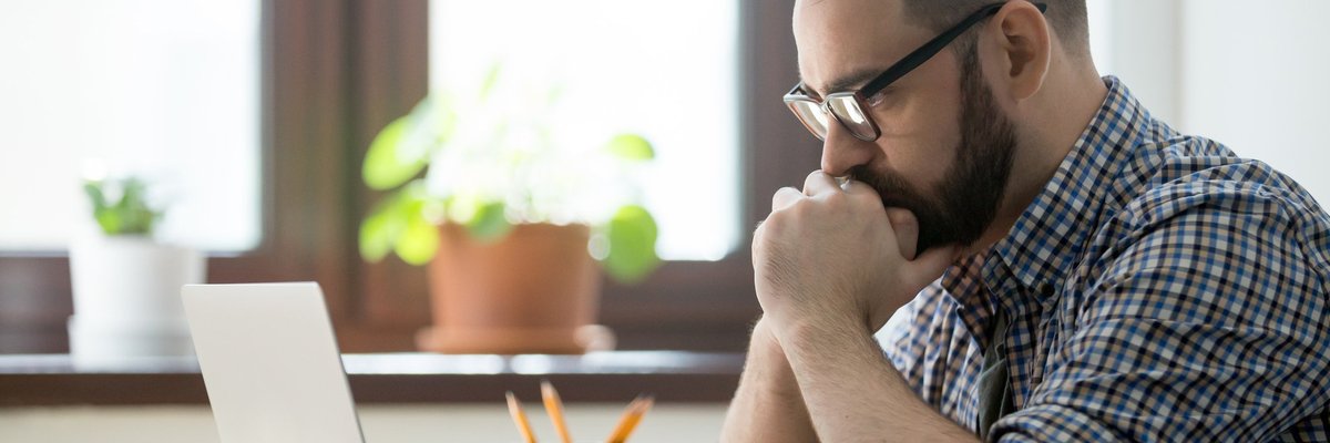 A stressed man sitting at a desk in front of an open laptop with his chin in his hands.