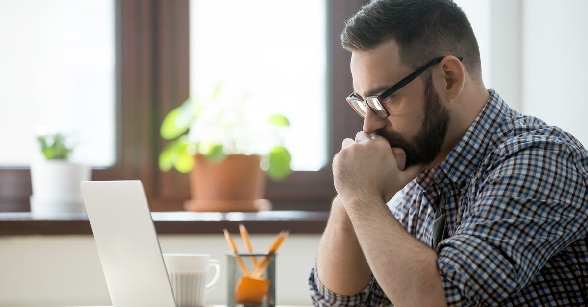 A man sitting at a desk with his laptop open and looking stressed with his chin in his hands.