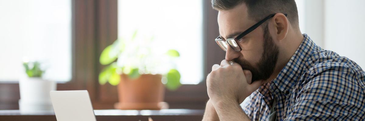 A man sitting at a desk with his laptop open and looking stressed with his chin in his hands.