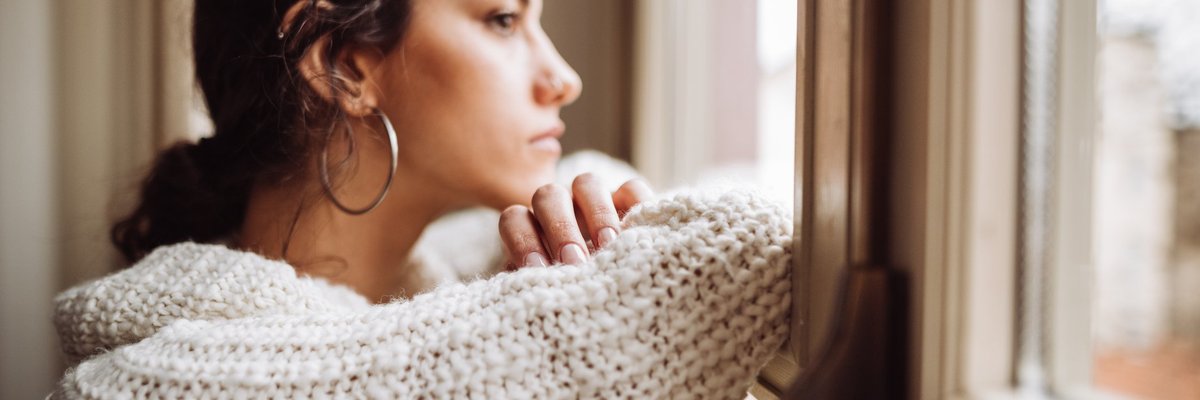 A stressed woman staring out the window with her head resting on her folded arms.
