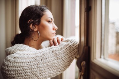 A stressed young woman looking out the window with her head resting on her crossed arms.