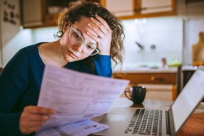 A stressed woman sitting in front of a laptop at her kitchen table and looking at bills in her hand.