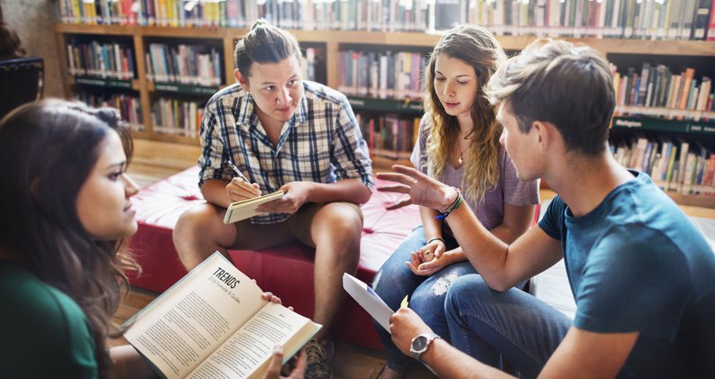 Group Of Students Talking In Library As One Takes Notes