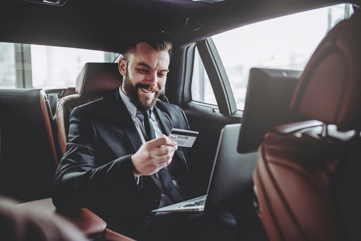 Man in suit in the back of a luxury car with laptop in his lap holding a credit card.