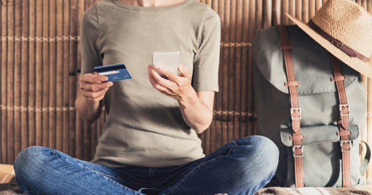 A woman holding a credit card and phone and sitting next to a backpack and hat.
