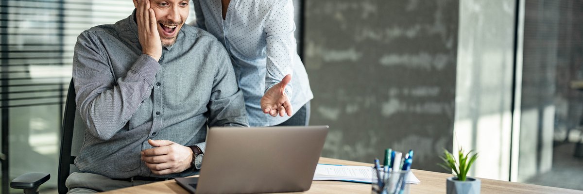 Two people in an office read the news on a computer and look pleasantly surprised.
