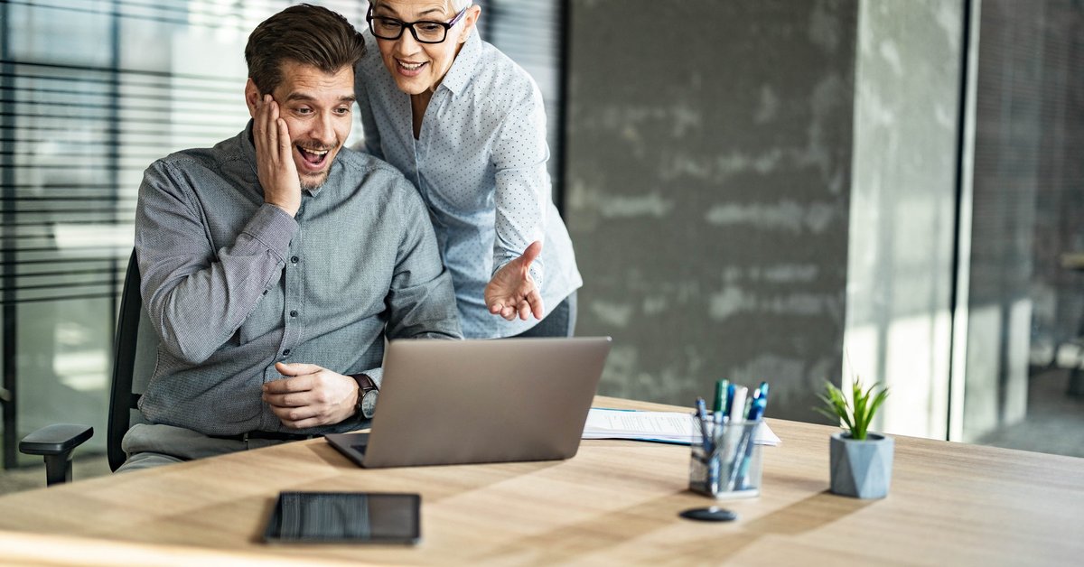 Two people in an office read the news on a computer and look pleasantly surprised.