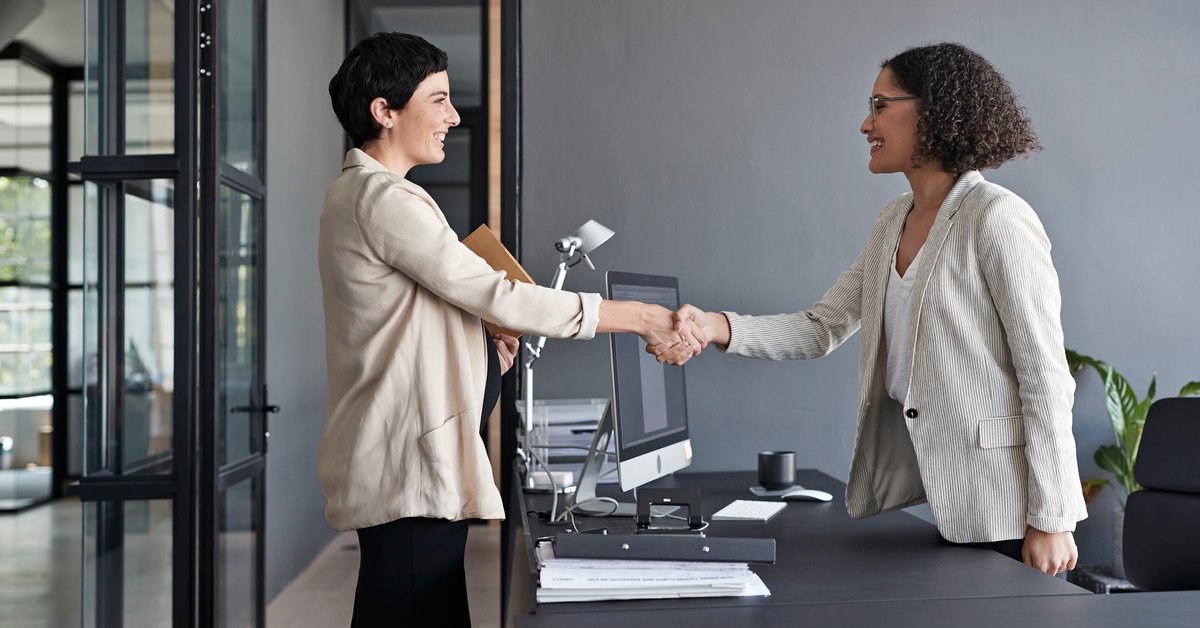 Two professionally dressed women shaking hands in an office