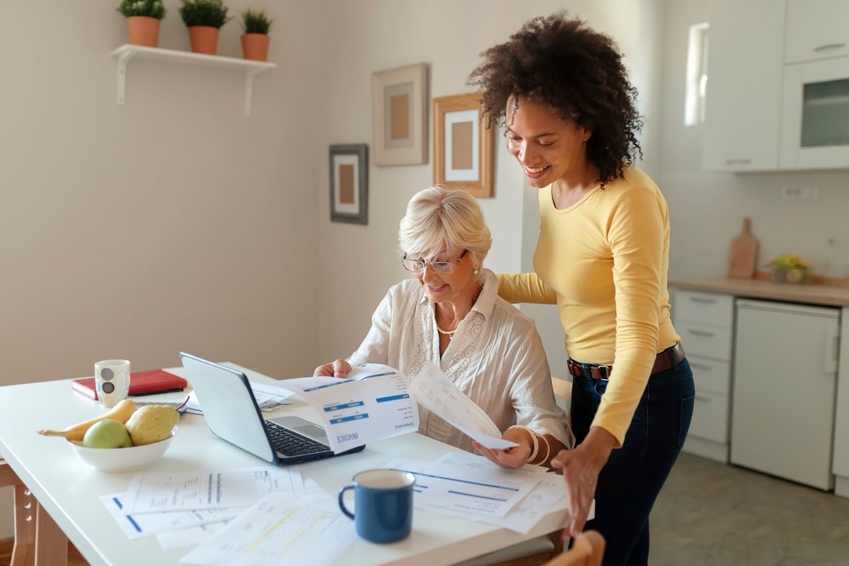 Two women are in a kitchen.  One sits as the other looks over her shoulder, helping with personal finances.
