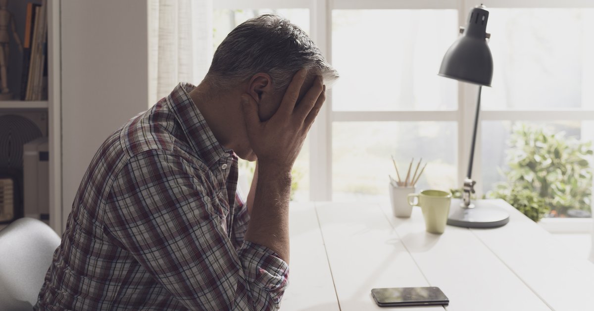 An upset man with his hands on his head on the phone at his desk.