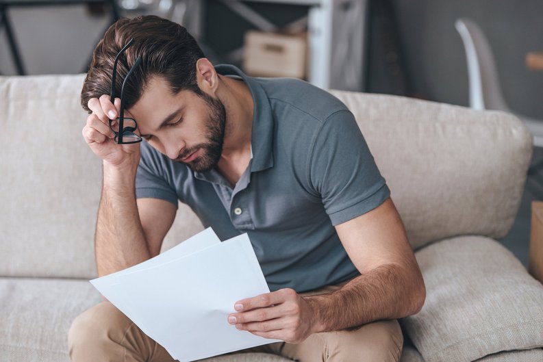 A tired-looking man resting his forehead on his hand while reading paperwork.