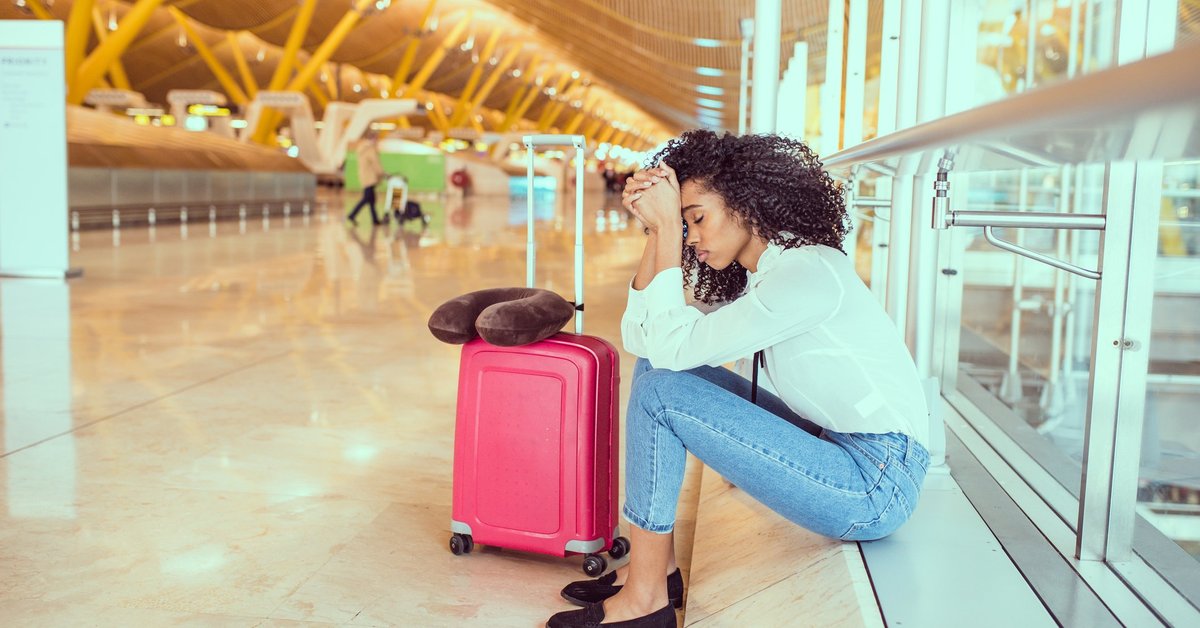 An upset woman at the airport sitting near a window next to her suitcase.