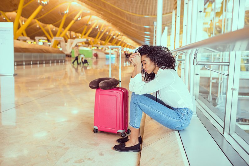 An upset woman at the airport sitting near a window next to her suitcase.