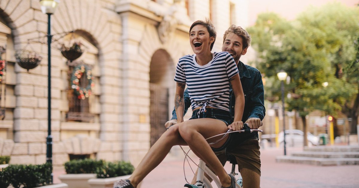 Young man riding a bike with a young woman laughing on the handlebars.