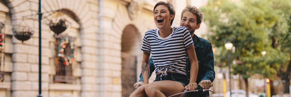 A laughing young woman riding on the handlebars of a bike being pedaled by a young man.