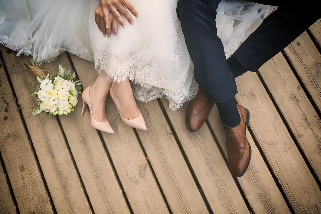 Wedding couple holding hands sitting on wooden deck beside bouquet.