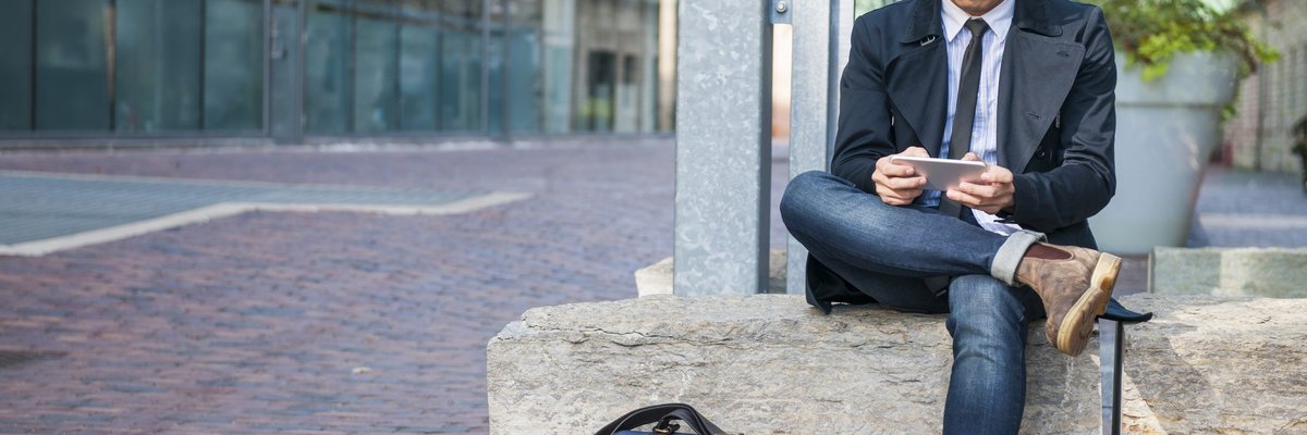 A well-dressed man on sitting on a stone bench and reading something on his phone.